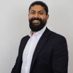 Professional headshot of Jyodh Bilan, a bearded man with dark hair slicked back, wearing a white dress shirt and a dark blazer. He is smiling and posed against a plain light background.