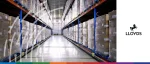 A long, brightly lit warehouse aisle featuring high industrial shelving stacked with numerous cardboard boxes wrapped in clear plastic. The Lloyds Bank logo, featuring their iconic black horse, is positioned on the right against a white background. A stylized, white digital wave pattern overlays the left side of the image, and a thin multi-colored banner runs along the bottom.