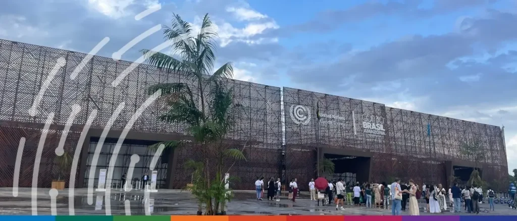 An outdoor wide shot of a large, modern pavilion with a facade made of dark, intricate latticework. The building features the United Nations Climate Change logo and text that reads "COP30 BRASIL AMAZÔNIA." Several people are walking or standing in the open plaza in front of the building under a cloudy blue sky. Palm trees are scattered near the entrance, and a stylized white fingerprint-like graphic is overlaid on the left side of the image. A colorful rainbow-striped banner runs along the bottom edge.