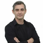 A professional studio headshot of Georgios Kolovos, a man with short, salt-and-pepper hair and a slight smile, wearing a black button-down shirt over a black t-shirt with his arms crossed against a solid white background.
