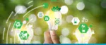 A hand holding a glowing lightbulb surrounded by green icons for recycling, renewable energy, and sustainable technology on a blurred nature background.