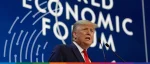 An image of US President Donald Trump speaking at the World Economic Forum (WEF) in Davos. He is wearing a dark suit and a bright red tie, standing behind a podium with two microphones. The background features a large blue banner with the "WORLD ECONOMIC FORUM" logo in white.
