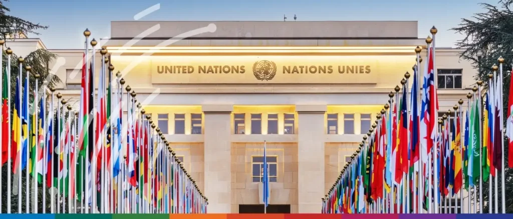 The building is a grand, symmetrical structure made of light-colored stone, prominently displaying the words "UNITED NATIONS" and "NATIONS UNIES" in gold lettering, flanking the UN emblem. In the foreground, two long, converging rows of diverse national flags on tall silver poles line a stone pathway leading toward the entrance. The scene is set under a clear sky, with the tops of green trees visible in the background, conveying a sense of global diplomacy and authority.